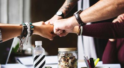 A group of friends fistbump over a table with a water bottle, a cup of pencils and pens, a laptop, and jar of granola.