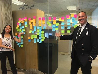 A content designer and a sharply-uniformed Fire Department Public Information Officer pose with a meeting room window covered in different colored stickies.