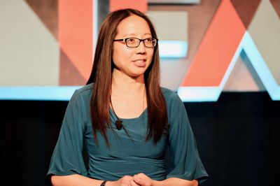 Asian woman with long hair speaks on a stage with a mic attached to her green long-sleeved shirt.